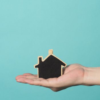 Small wood house in hands represent concepts such as home care family love real estate housing shelter insurance and mortgage. Hands holding small model house isolated on blue green studio background.